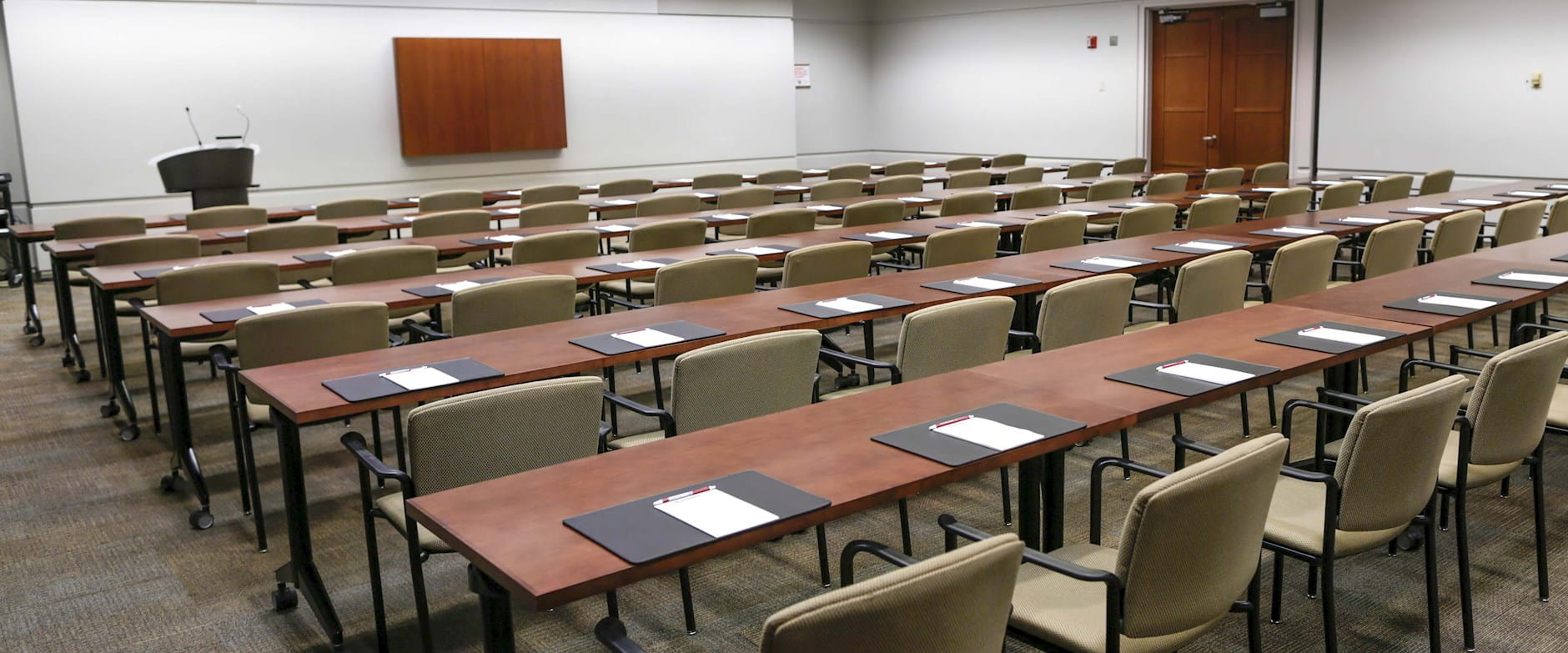 View from the back of a meeting room with rows of tables and chairs and a podium at the front