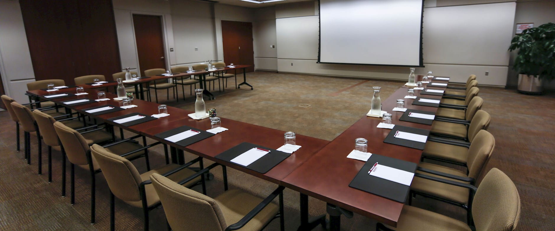 Angled view of warm wood tables in a U-shape facing a projector screen with a green potted plant in the corner