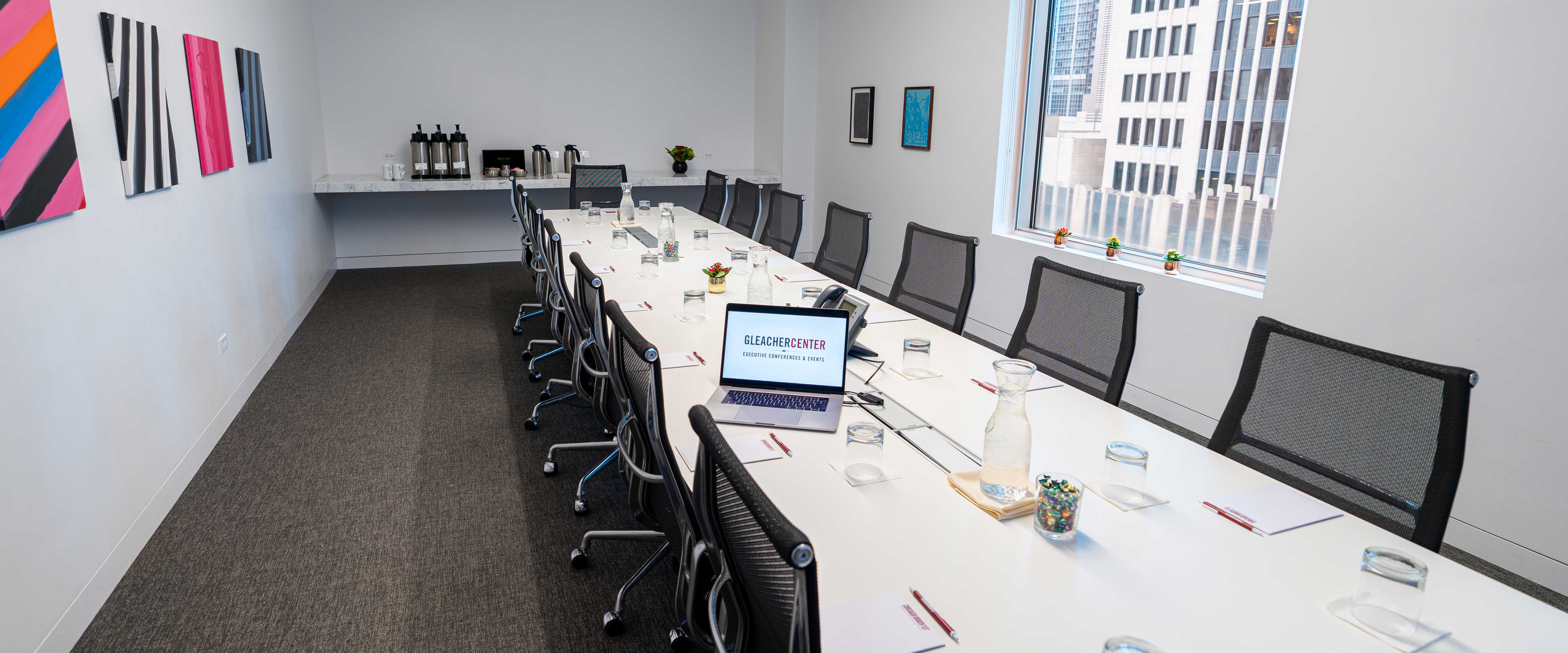 Boardroom with paintings, coffee station, and a long table set with notepads and a laptop in the Gleacher Center