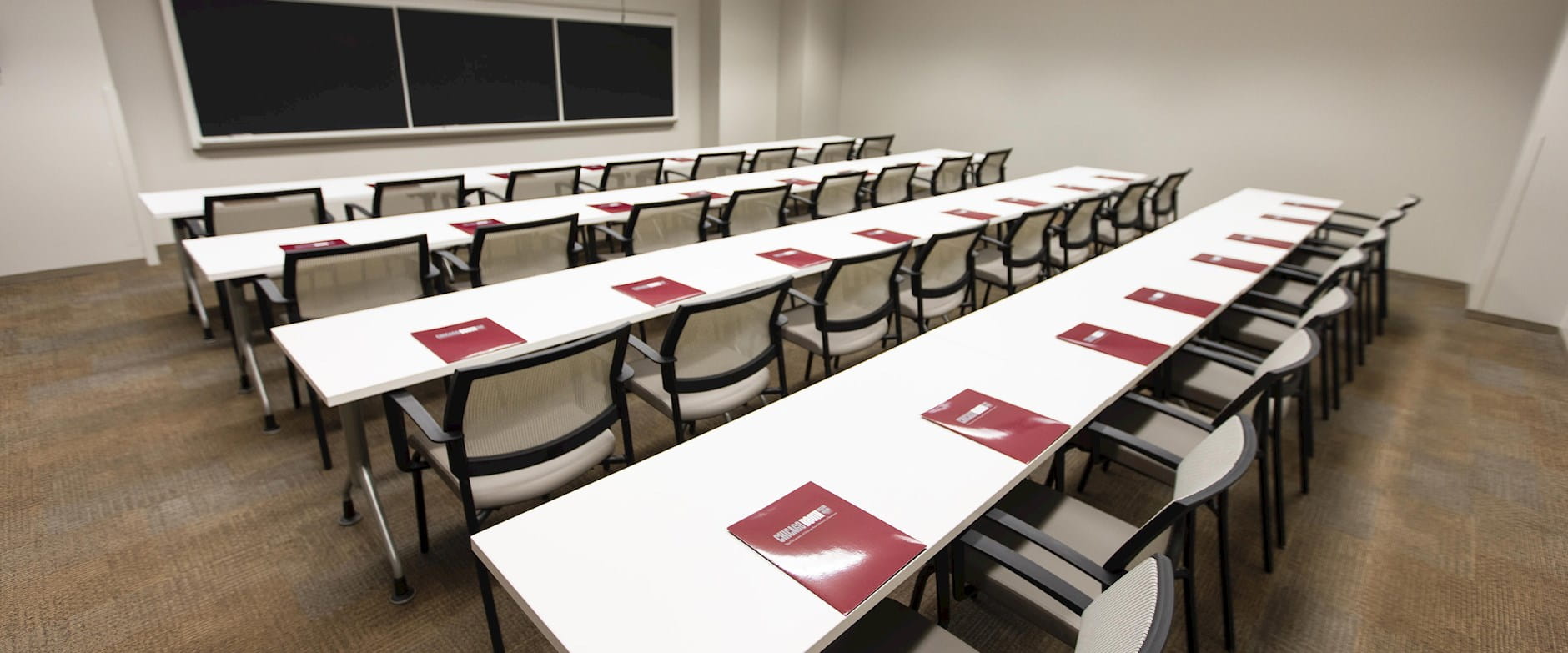 Classroom 203 at the University of Chicago Gleacher Center from the entrance with 3 rows of tables and chairs in front of a blackboard