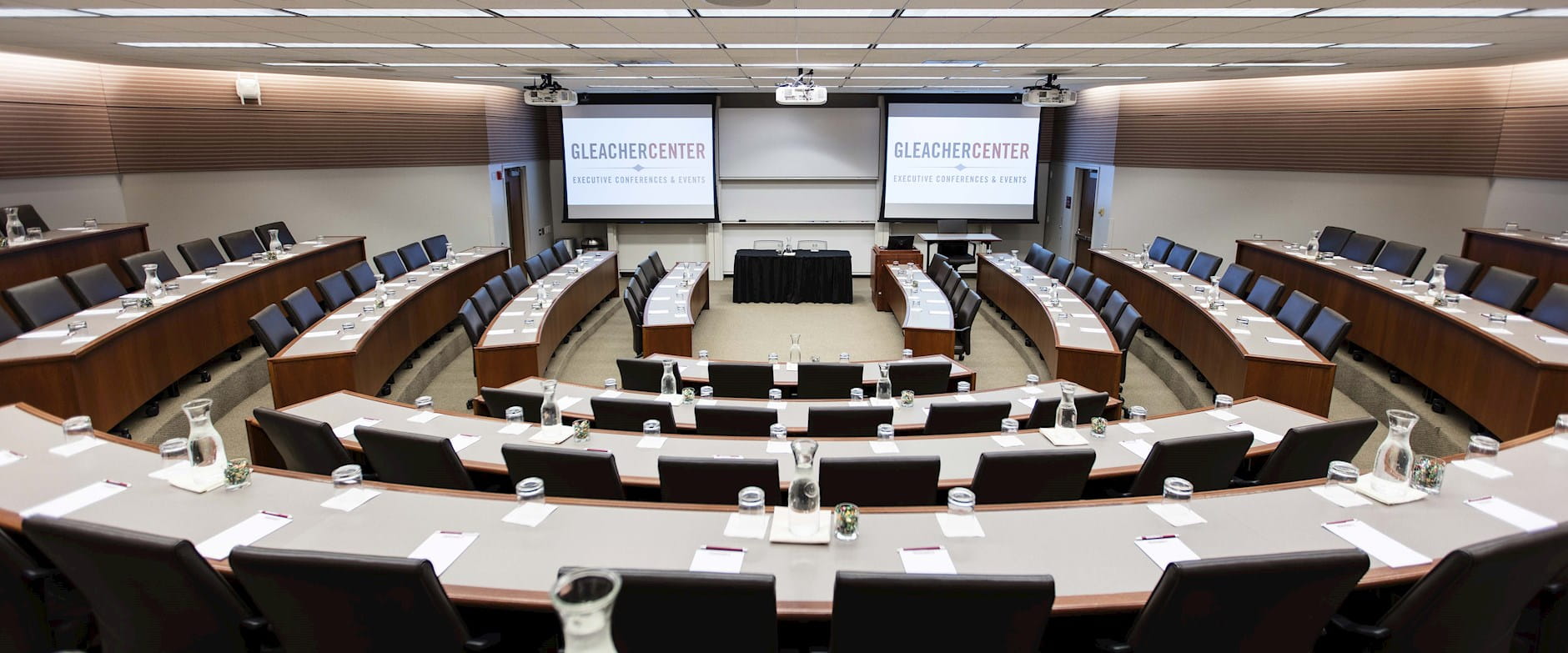 View from the back of room 400 at the Gleacher Center looking out over tiered tables and chairs in a semi-circle with a presenters' table and 2 projector screens at the front
