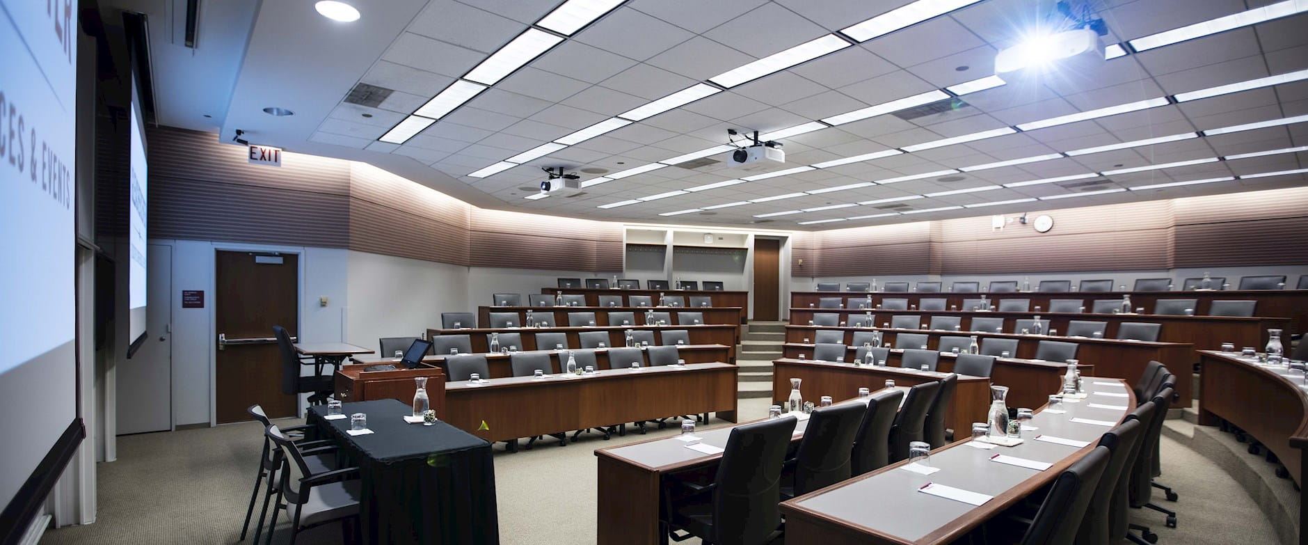 A wide view from the front-left of meeting room 400 at the Gleacher Center looking at tiered tables and chairs in a semi-circle; a projector shines brightly from the ceiling